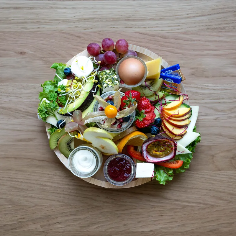 Vegetarische Frühstücksplatte mit Käse, Ei, Avocado und Obst auf einem runden Holzbrett